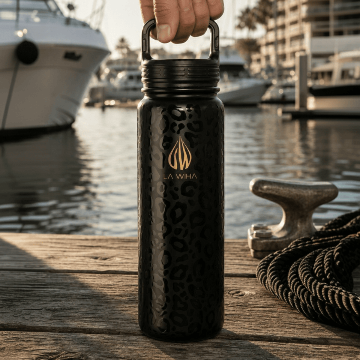 A 24 ounce black insulated water bottle with a gold logo and leopard-style pattern, held by a hand on a wooden dock marina port with luxury high end boats and water in the background.