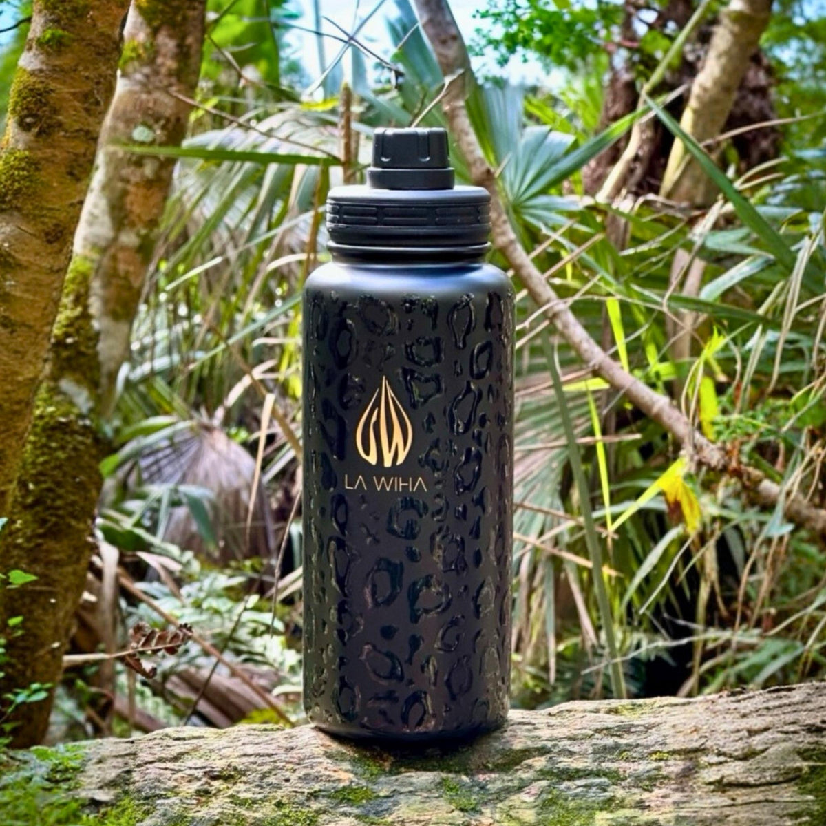 A durable 32 ounce black water bottle with a gold La Wiha logo, sitting on a mossy log in a lush green forest. hunting, fishing, walking, hiking,