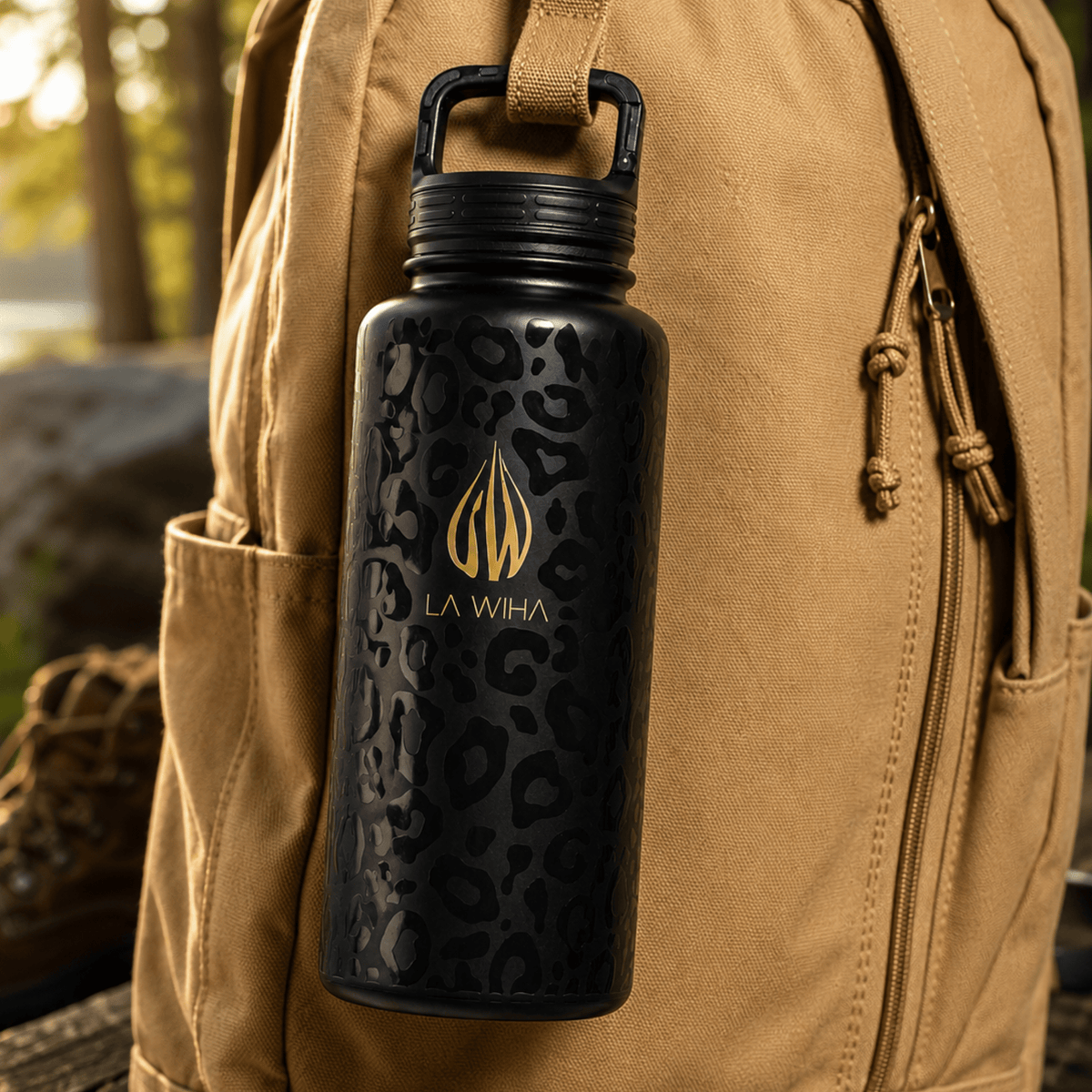 A 32 ounce stainless steel black insulated water bottle with a gold La Wiha logo and dark leopard print, clipped by the built in carabiner clip lid hanging from a tan hiking backpack outdoors, lake front, waterfront, hiking boot in the background, log, trees, water