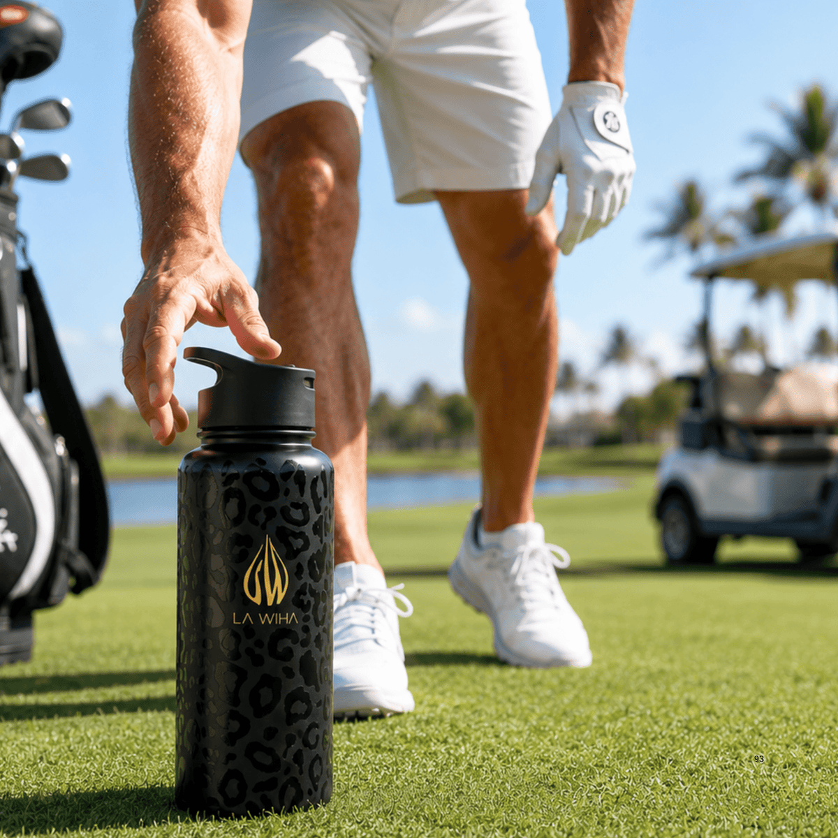 A close-up of a black water bottle with a gold logo, held by a golfer on a green golf course. The background shows a blurred golf cart, palm trees, blue sky, golf course, golf bag, pond