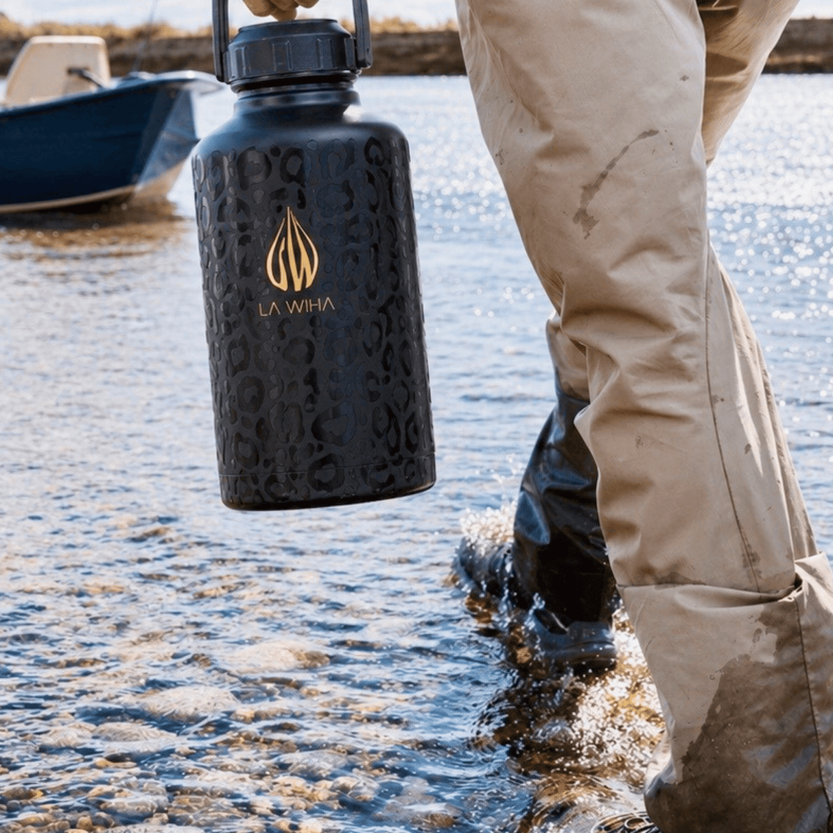 A person in beige waders and black boots stands in shallow water, pond, lake, water, fishing, fishing boat holding a large black insulated water bottle with a gold logo and the text 'LA WIHA' on the front.