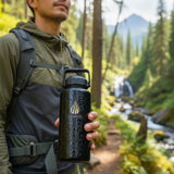 A man hiking walking outdoors in a forest, next to a river, stream ,mountains, outdoors, while holding a 32 ounce black La Wiha water bottle with a gold logo. The background shows a blurred stream, trees, walking path and mountains.