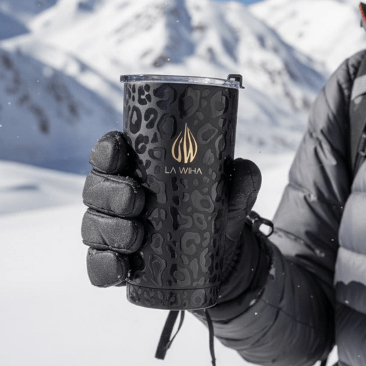 A person in a snowy mountains, skiing, snowboarding, playing in the snow, holding a black insulated tumbler with a tonal leopard pattern and a gold LA WIHA logo. The person is wearing black gloves and a gray puffer jacket. cabin