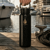 A 24 ounce black insulated water bottle with a gold logo and leopard-style pattern, held by a hand on a wooden dock marina port with luxury high end boats and water in the background.