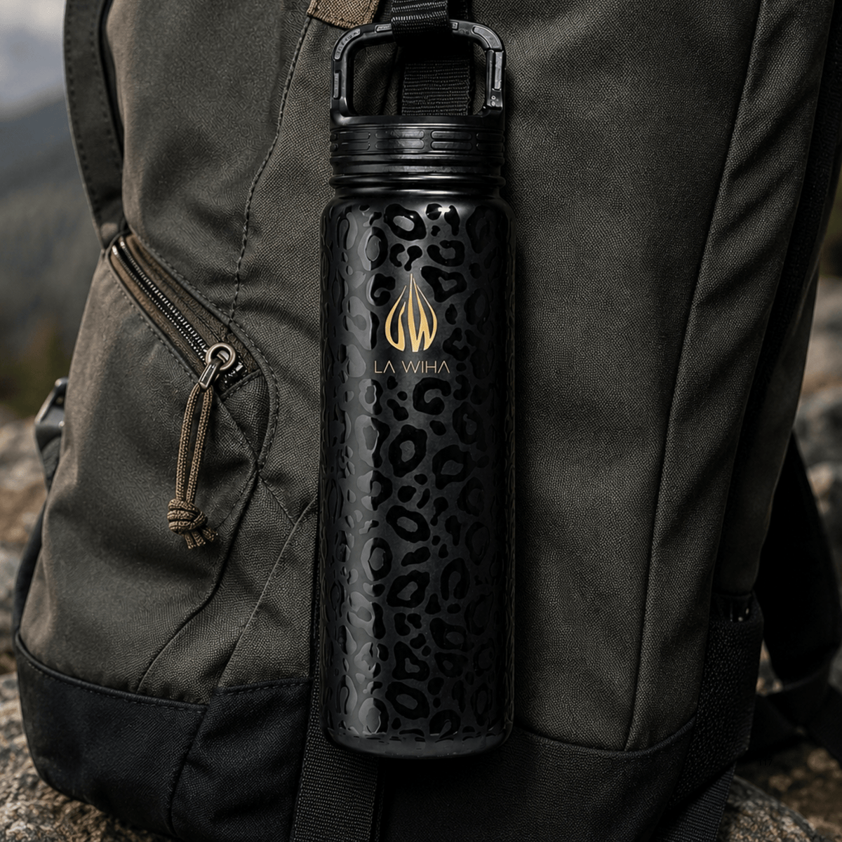 A 24 ounce leak proof black insulated water bottle with a leopard print pattern and gold LA WIHA logo, clipped and hanging from a dark gray backpack outdoors  mountain, hiking, back packing, adventure, extreme sport, rock climbing