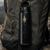 A 24 ounce leak proof black insulated water bottle with a leopard print pattern and gold LA WIHA logo, clipped and hanging from a dark gray backpack outdoors  mountain, hiking, back packing, adventure, extreme sport, rock climbing