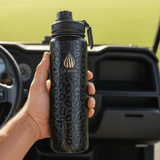 A hand holds a 20 ounce black insulated water bottle with a subtle leopard print pattern and gold 'LA WIHA' branding, set against the blurred background of a golf cart interior. golfing golf putt putt green golf course