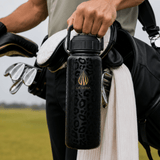A close-up of a man on a luxury lush golf course holding a black 24 ounce stainless steel insulated water bottle with a gold logo and leopard print. The person is also carrying a black golf bag and holding golf clubs on a golf course