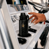 A black insulated water bottle with a subtle leopard print pattern and gold 'LA WIHA' branding, resting in a cup holder on a boat's helm station while the captain's hand reaches toward it. deep sea fishing, yacht, boat, fishing, yachting, lake day, seaside, ocean, sea