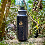 A durable 32 ounce black water bottle with a gold La Wiha logo, sitting on a mossy log in a lush green forest. hunting, fishing, walking, hiking, 