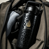 A 32 ounce black stainless steel La Wiha water bottle with a gold logo and leopard print pattern, shown inside an open olive green backpack, workbag, gym bag, suitcase, duffel bag, airport , travel, traveler 