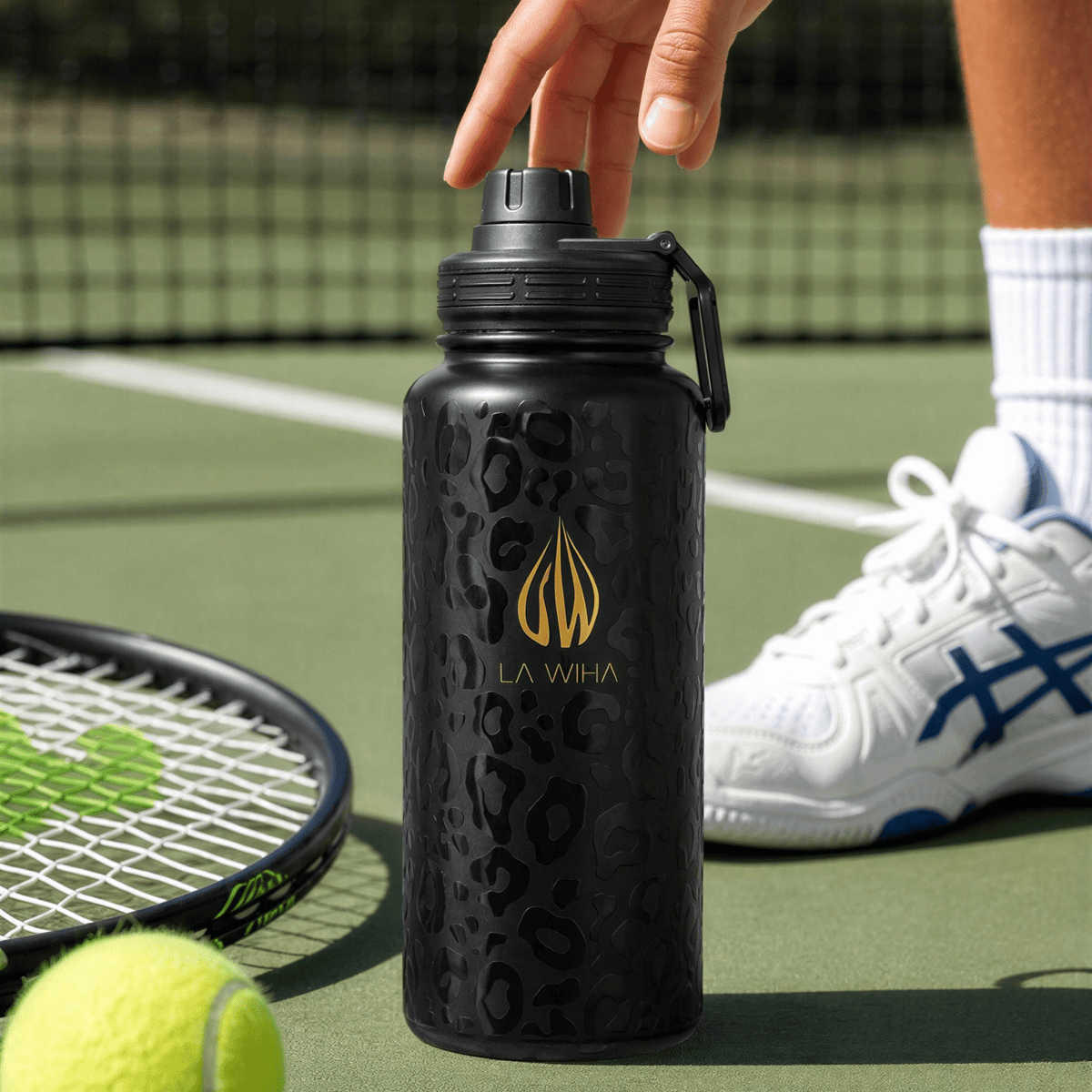 A 32 ounce black water bottle with a gold 'LA WIHA' logo and leopard print pattern, with a sport lid and a hand near a green tennis court with a tennis racket, tennis ball, and a player's tennis shoe visible on a green clay  tennis court, athlete, leisure, all day hydration, keeps cold in the heat