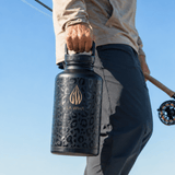 A person outdoors holding a large black LA WIHA durable larger half gallon insulated bottle with a gold logo . The person is wearing a beige long-sleeve shirt and dark pants against a clear blue sky, and a fishing rod is visible.