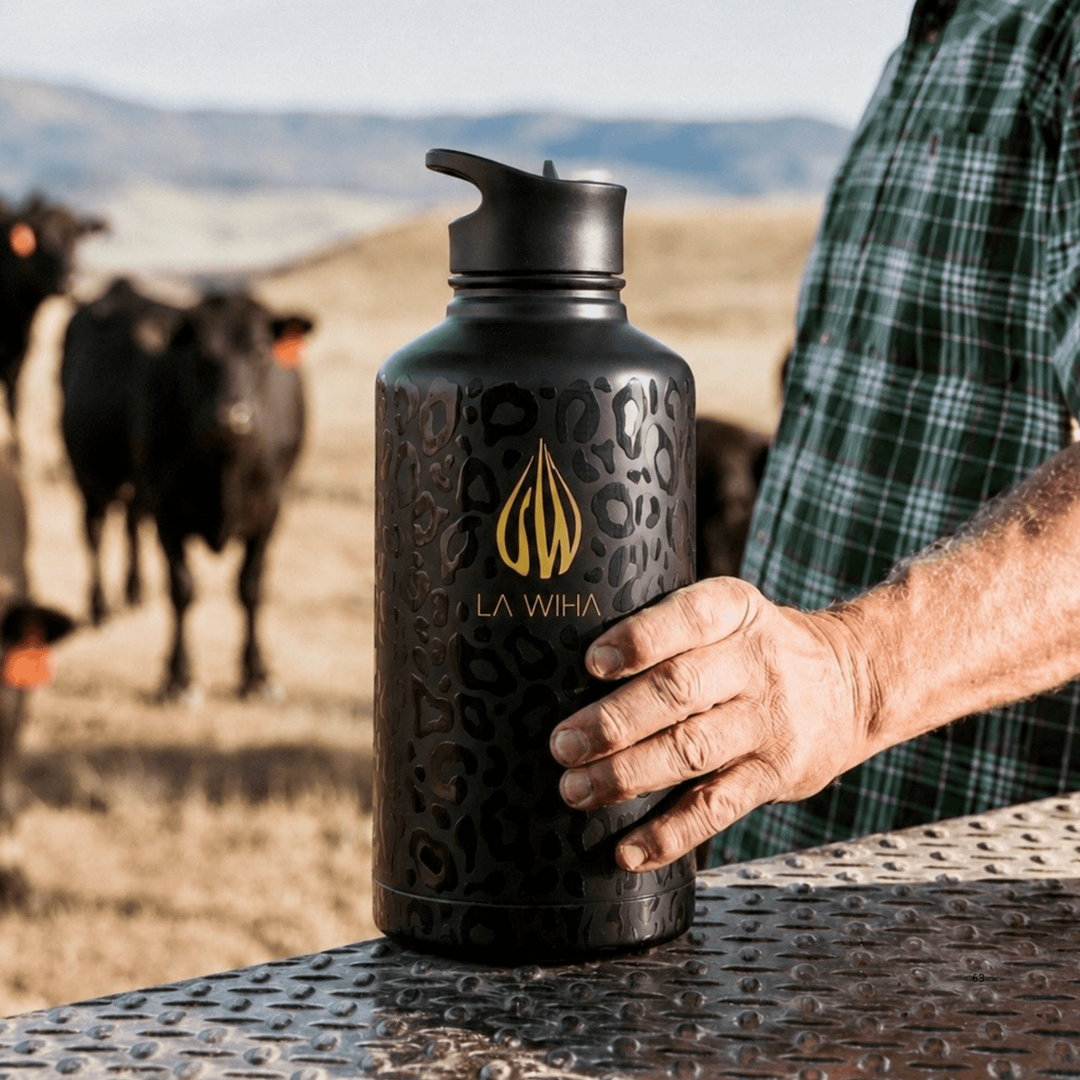 A person in a green plaid shirt holds a large black La Wiha water jug with a gold logo and subtle leopard pattern, set against a blurred outdoor ranch background with cows, back of a truck on a truck bed, rancher, suitable for out doors