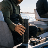 A man in fishing waders and an olive green long-sleeve shirt holds a black insulated travel tumbler with a gold logo while sitting in a boat. A fly fishing rod and reel rest on the boat next to him, with water, shoreline, lake, river, bank visible in the background.