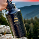 Black 64oz half gallon water bottle with tan logo and text held by a hand against a mountainous background
